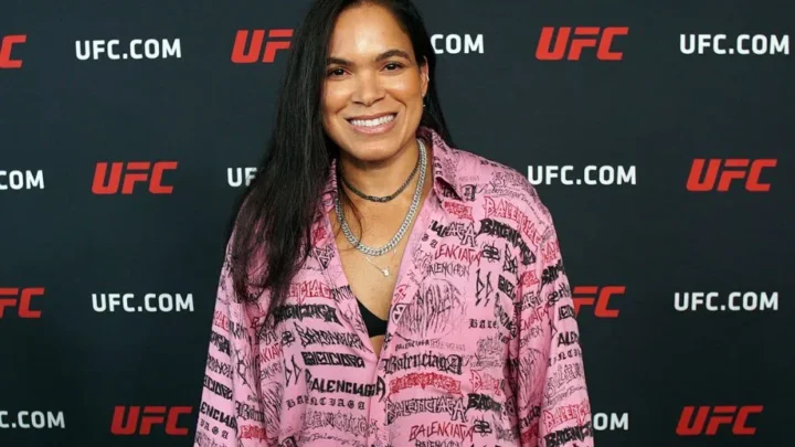Mulher sorridente, de cabelo longo e escuro, posa em frente a painel com logos do UFC usando camisa rosa estampada com letras pretas e vermelhas, além de colares prateados