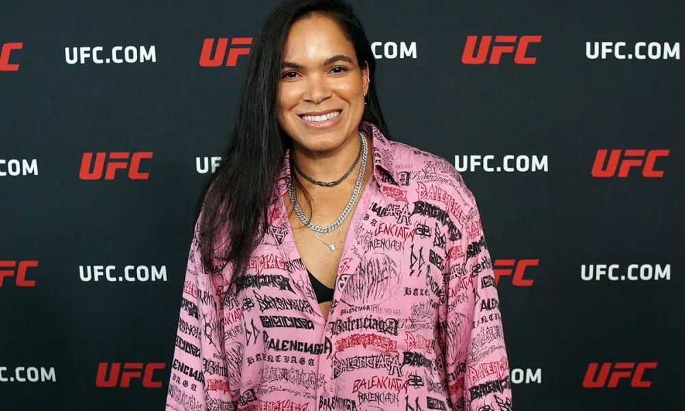 Mulher sorridente, de cabelo longo e escuro, posa em frente a painel com logos do UFC usando camisa rosa estampada com letras pretas e vermelhas, além de colares prateados