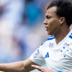 Jogador de camisa branca com detalhes azuis e estrelas do Cruzeiro gesticula com a mão aberta em campo