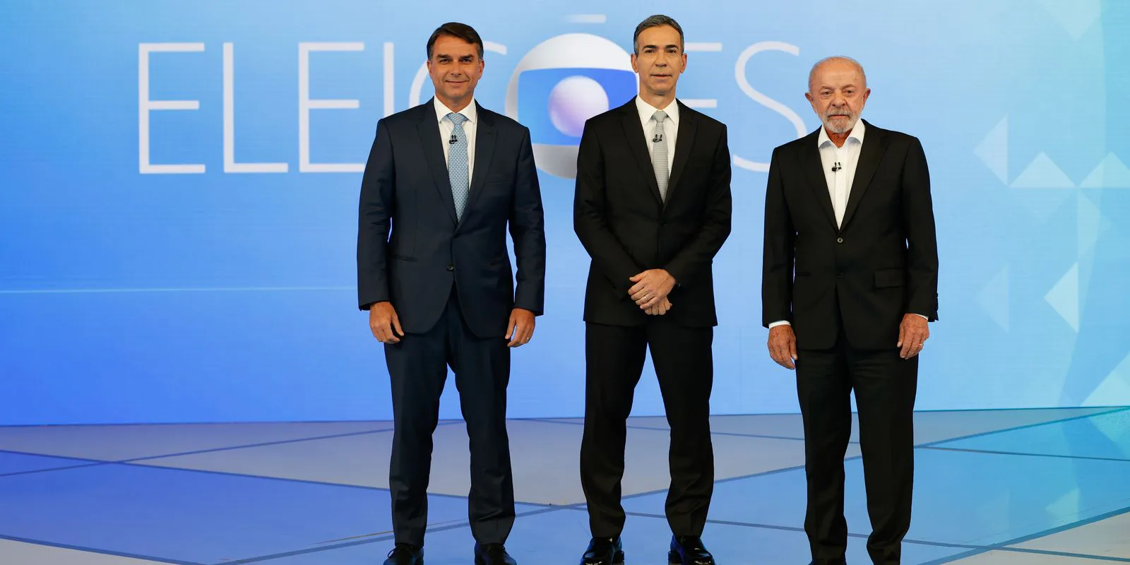 Flávio Bolsonaro, o mediador César Tralli e Lula, de corpo inteiro, no cenário do debate da TV Globo, no Rio de Janeiro, em 23 de outubro de 2026