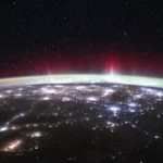 Aurora verde e vermelha sobre a curvatura da Terra, acima das luzes das cidades da Europa e da Rússia, fotografada da Estação Espacial em fevereiro de 2026