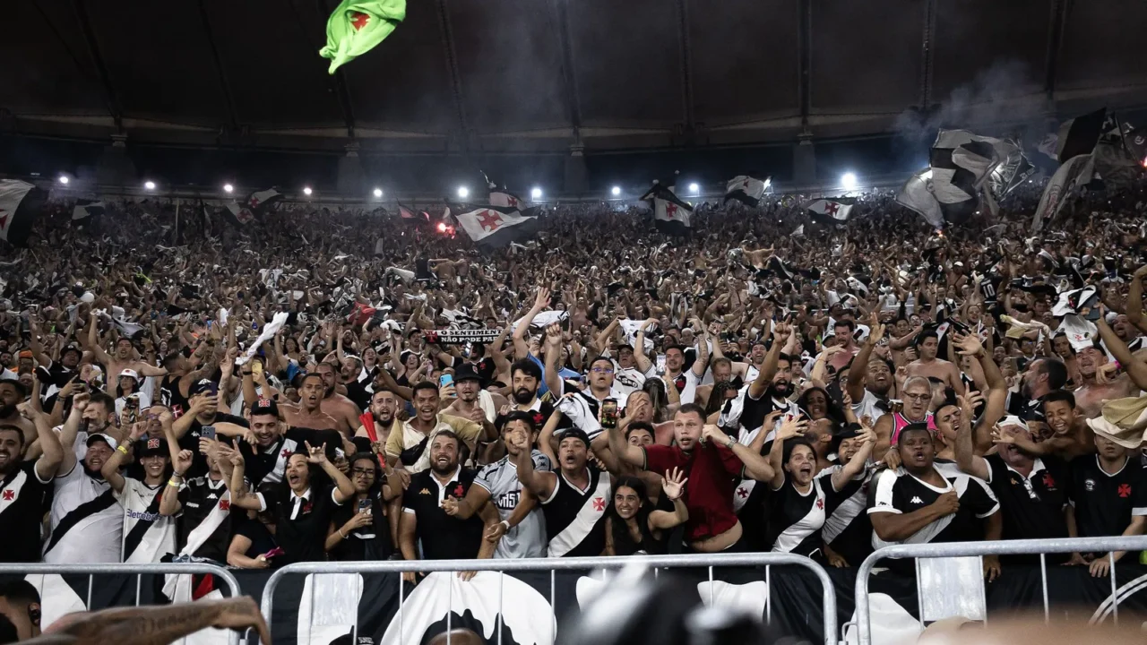 Torcedores do Vasco, de preto e branco, comemoram e erguem bandeiras e braços em um estádio lotado, com fumaça ao fundo
