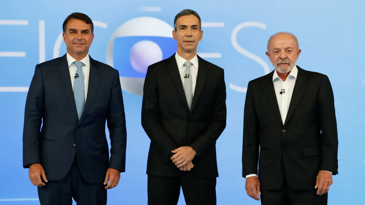 Flávio Bolsonaro, à esquerda, de terno azul-escuro e gravata azul-clara, e Lula, à direita, de terno preto e camisa branca, de pé diante do painel "Eleições" no estúdio do debate da TV Globo, com um homem de terno preto entre os dois, no Rio de Janeiro, em 23 de outubro de 2026