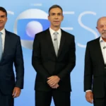 Flávio Bolsonaro, à esquerda, de terno azul-escuro e gravata azul-clara, e Lula, à direita, de terno preto e camisa branca, de pé diante do painel "Eleições" no estúdio do debate da TV Globo, com um homem de terno preto entre os dois, no Rio de Janeiro, em 23 de outubro de 2026