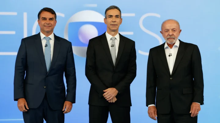 Flávio Bolsonaro, à esquerda, de terno azul-escuro e gravata azul-clara, e Lula, à direita, de terno preto e camisa branca, de pé diante do painel "Eleições" no estúdio do debate da TV Globo, com um homem de terno preto entre os dois, no Rio de Janeiro, em 23 de outubro de 2026