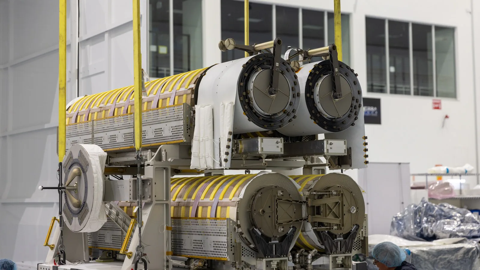 Técnicos acompanham um guindaste que baixa o par de painéis solares enrolados, cobertos de faixas amarelas, numa sala limpa do Centro Espacial Kennedy