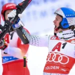 Esquiador de macacão e capacete azul, com braço erguido segurando um esqui vermelho, gritando de alegria em pista de neve, ao lado de outro atleta de vermelho ao fundo