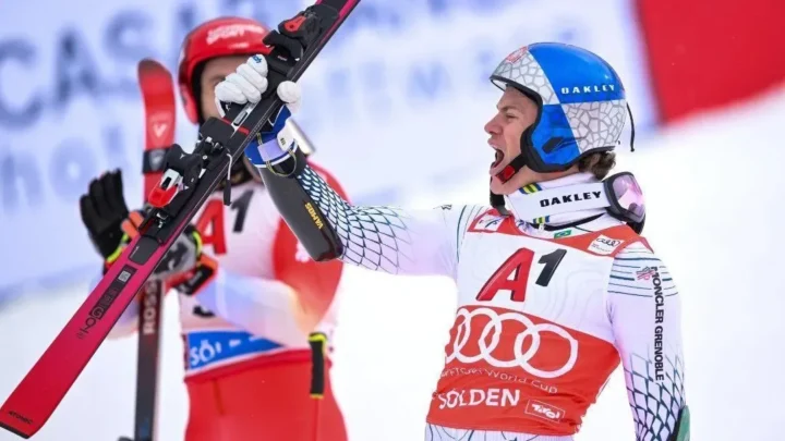 Esquiador de macacão e capacete azul, com braço erguido segurando um esqui vermelho, gritando de alegria em pista de neve, ao lado de outro atleta de vermelho ao fundo