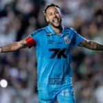 Jogador de camisa azul do Santos, com faixa de capitão no braço, comemora de braços abertos e sorrindo em um estádio