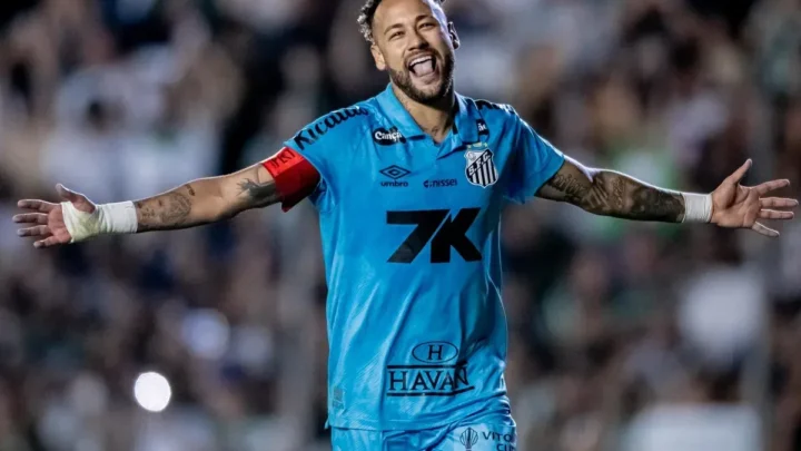 Jogador de camisa azul do Santos, com faixa de capitão no braço, comemora de braços abertos e sorrindo em um estádio