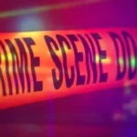 Fita de isolamento com a inscrição "Crime Scene Do Not Cross" em close, com iluminação vermelha e roxa de fundo desfocado
