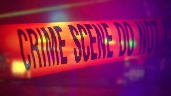 Fita de isolamento com a inscrição "Crime Scene Do Not Cross" em close, com iluminação vermelha e roxa de fundo desfocado