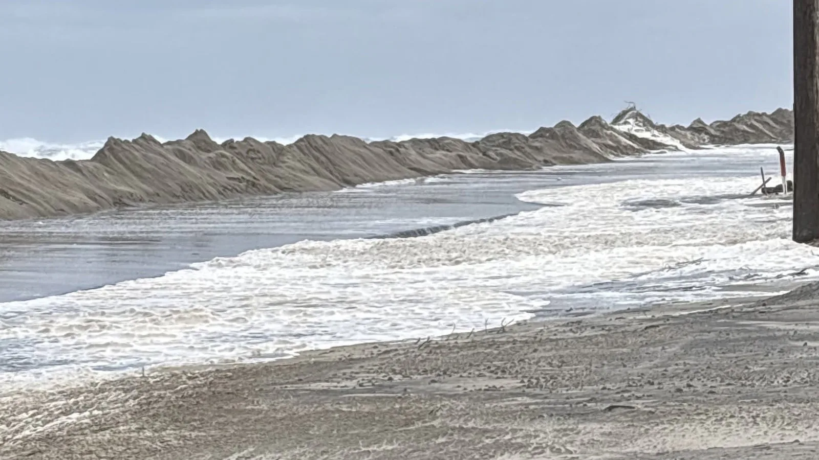 Ondas atravessam a faixa de areia e chegam à beira da estrada, com montes de areia empurrados pelo mar ao fundo