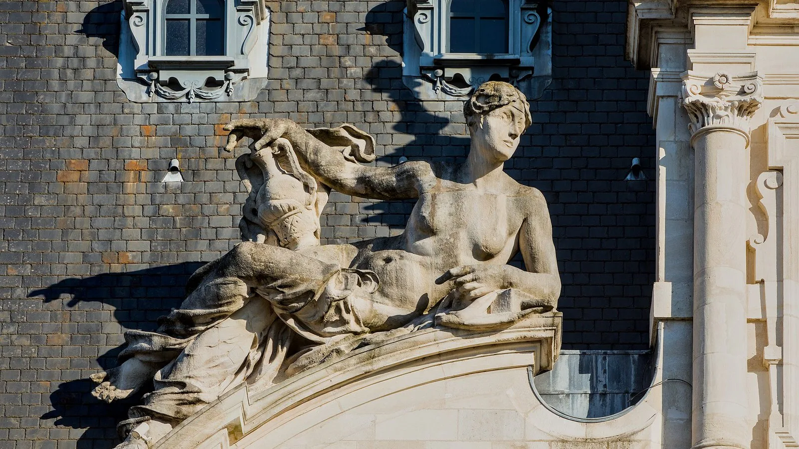 Escultura de figura feminina reclinada sobre o frontão da prefeitura de Limoges, diante do telhado de ardósia, em maio de 2026