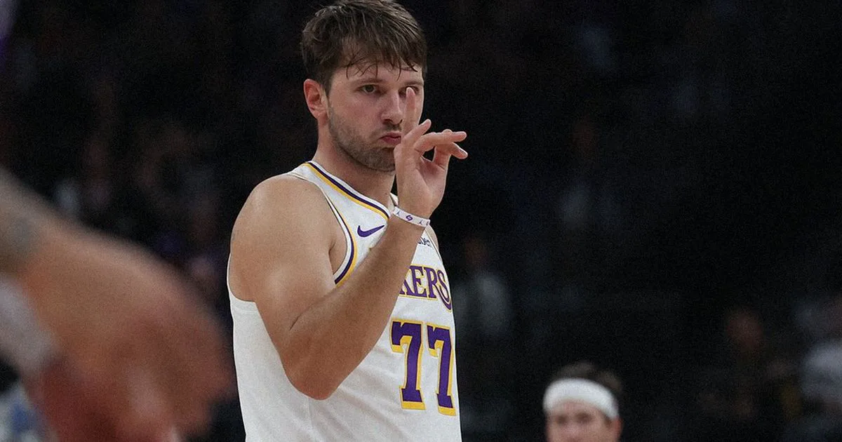 Jogador de camisa branca do Lakers, número 77, faz gesto com a mão tocando o nariz durante partida em quadra com torcida ao fundo