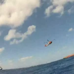 Helicóptero da Guarda Costeira sobre o mar, visto da água, com embarcações no horizonte