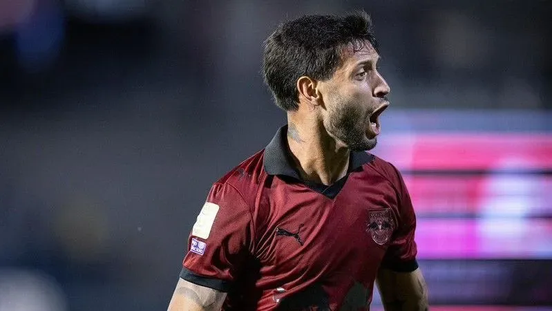 Jogador com camisa vinho e preta da Puma, gritando em campo com estádio desfocado ao fundo