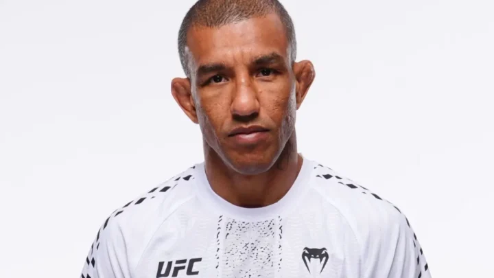 Atleta de UFC em retrato frontal contra fundo branco, vestindo uniforme branco da organização com logotipos em preto