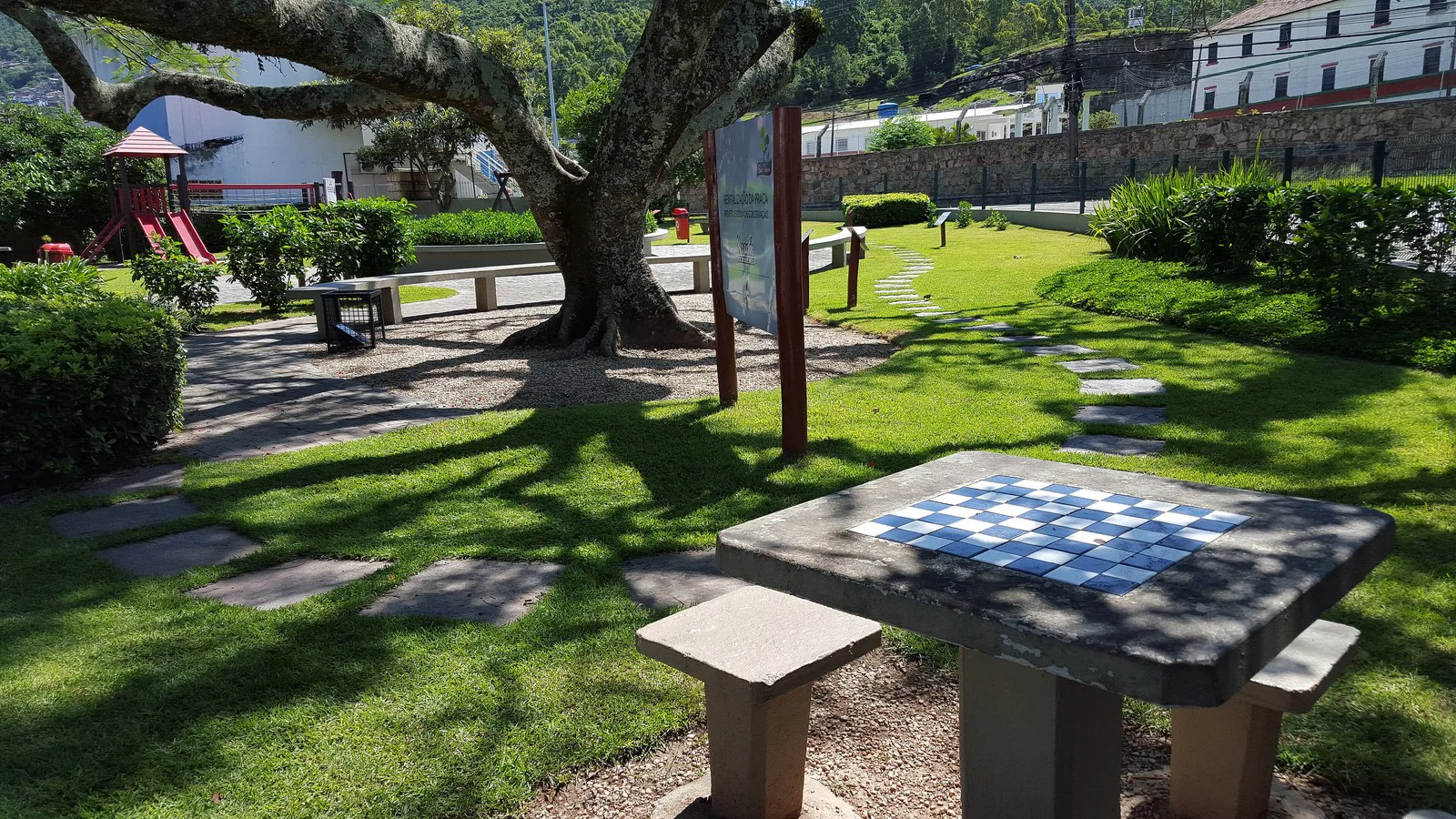 Mesa de concreto com tabuleiro de xadrez azul e branco pintado no tampo e dois bancos, em um parque público arborizado (imagem ilustrativa)