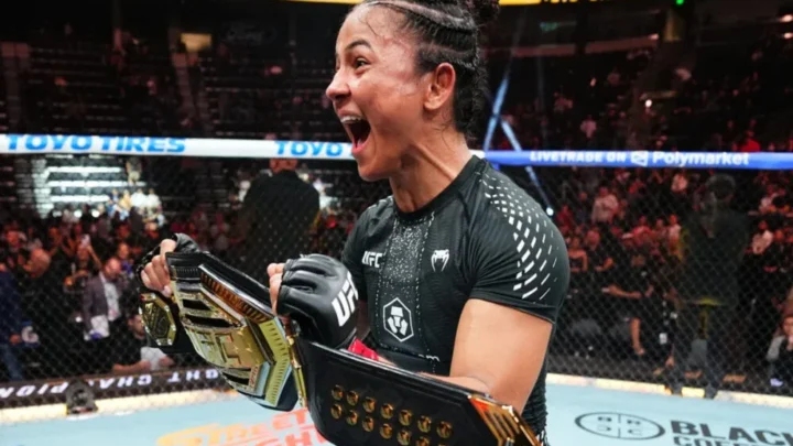 Lutadora de MMA com cinturão dourado do UFC comemora de punhos cerrados e boca aberta dentro do octógono, cercada por torcida nas arquibancadas