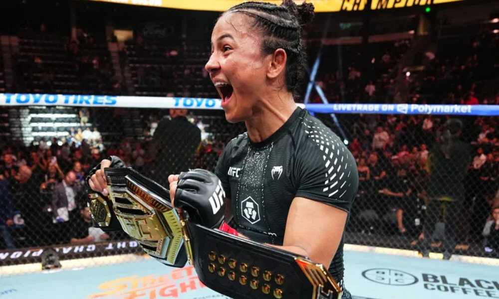 Lutadora de MMA com cinturão dourado do UFC comemora de punhos cerrados e boca aberta dentro do octógono, cercada por torcida nas arquibancadas
