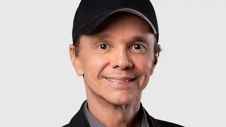Homem de boné preto e jaqueta escura, sorrindo, em foto de fundo cinza claro