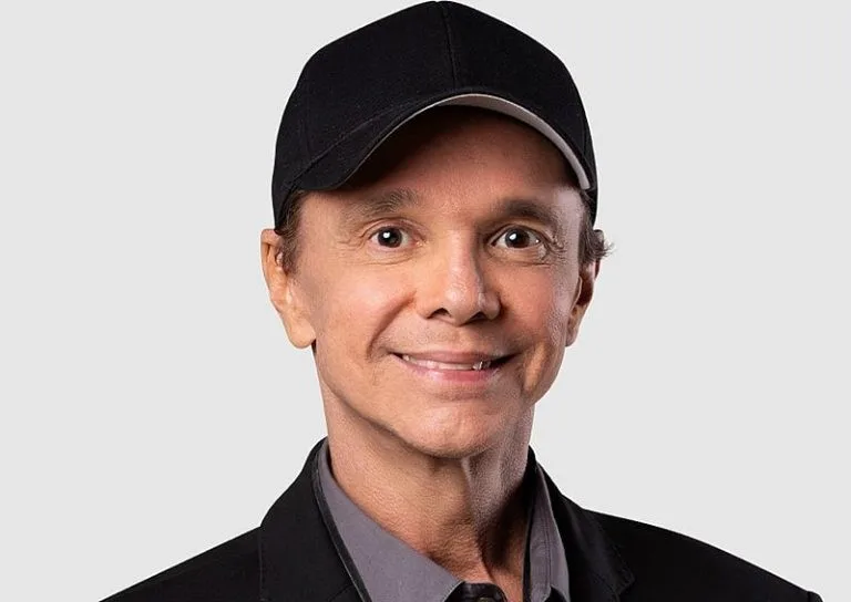 Homem de boné preto e jaqueta escura, sorrindo, em foto de fundo cinza claro