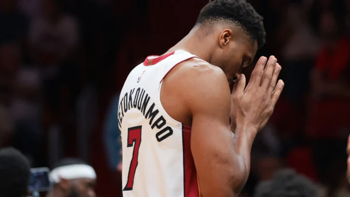 Jogador de basquete com camisa branca e vermelha, número 7 e sobrenome Antetokounmpo nas costas, leva as mãos juntas ao rosto durante partida em quadra