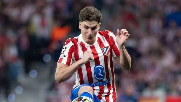 Jogador de camisa listrada vermelha e branca, com escudo do clube e patrocínio da Riyadh Air, chuta a bola durante partida em estádio lotado