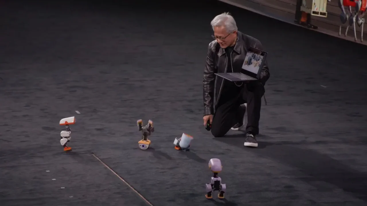 Jensen Huang, CEO da Nvidia, ajoelhado no palco com um notebook na mão enquanto um dos robôs-pato Microduck fica de cabeça para baixo ao dar uma cambalhota, na keynote da GTC Berlim, em 21 de outubro de 2026