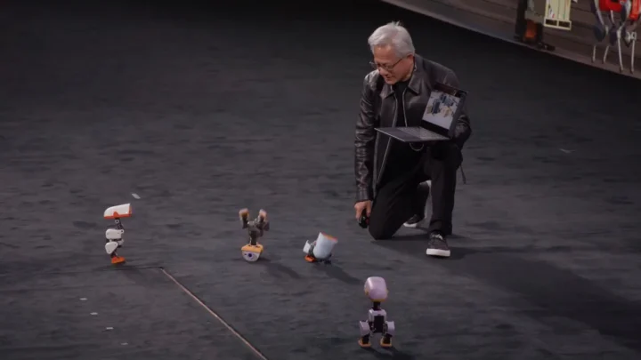 Jensen Huang, CEO da Nvidia, ajoelhado no palco com um notebook na mão enquanto um dos robôs-pato Microduck fica de cabeça para baixo ao dar uma cambalhota, na keynote da GTC Berlim, em 21 de outubro de 2026