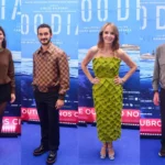 Quatro convidados posam em tapete azul com cartaz do filme "100 Dias", da esquerda para a direita: uma mulher de suéter marrom e minissaia preta, um homem de camisa xadrez marrom, uma mulher de vestido verde texturizado e um homem de jaqueta cinza