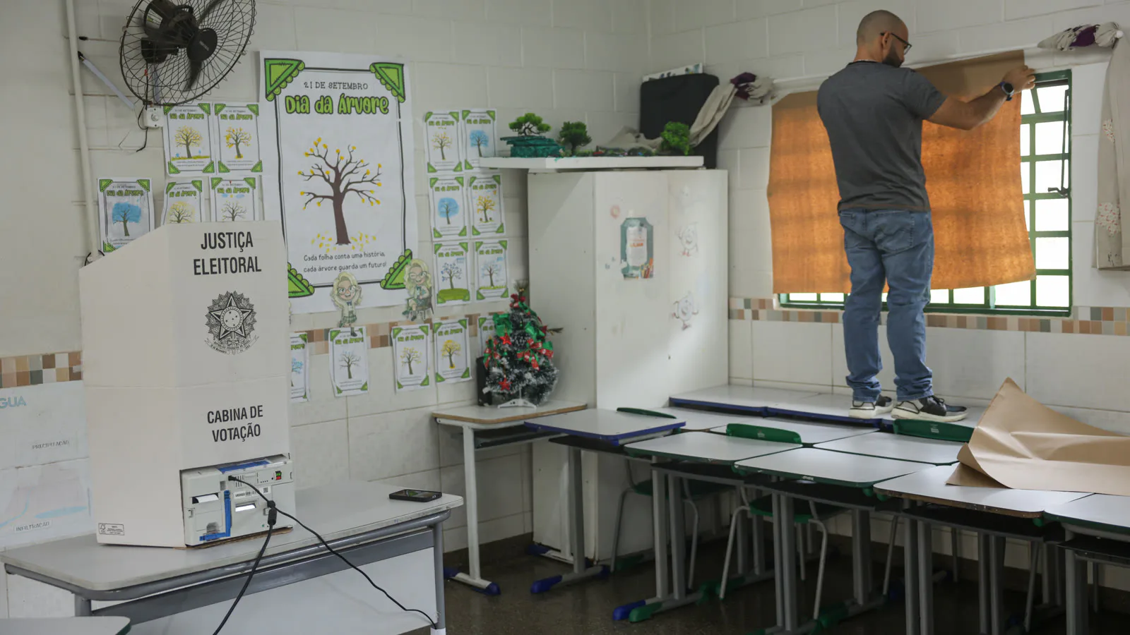 Cabine de votação da Justiça Eleitoral montada sobre uma carteira escolar enquanto um homem cobre a janela da sala com papel pardo, em Brasília, em 24 de outubro de 2026