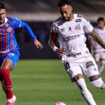 Jogador de camisa branca com a número 10 e braçadeira de capitão conduz a bola em campo, sendo marcado de perto por adversário de uniforme azul e vermelho
