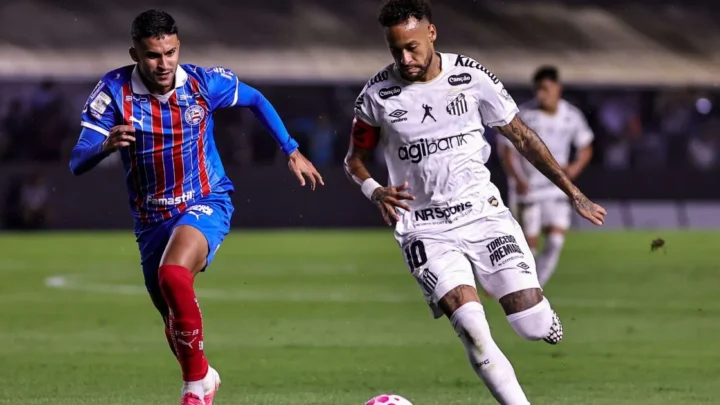 Jogador de camisa branca com a número 10 e braçadeira de capitão conduz a bola em campo, sendo marcado de perto por adversário de uniforme azul e vermelho