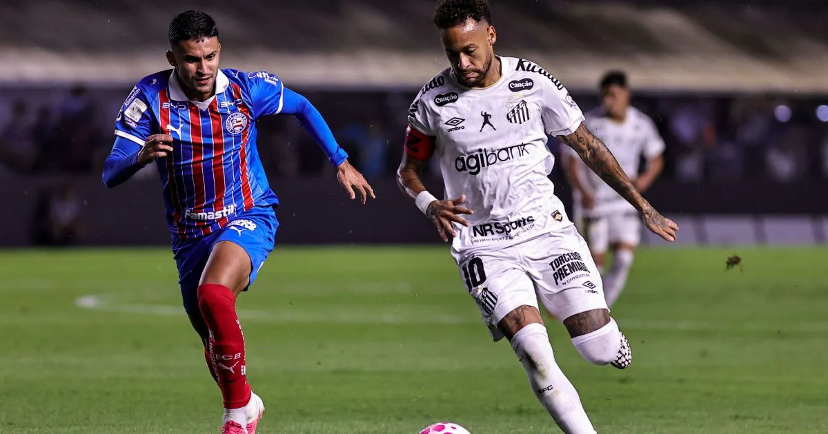 Jogador de camisa branca com a número 10 e braçadeira de capitão conduz a bola em campo, sendo marcado de perto por adversário de uniforme azul e vermelho