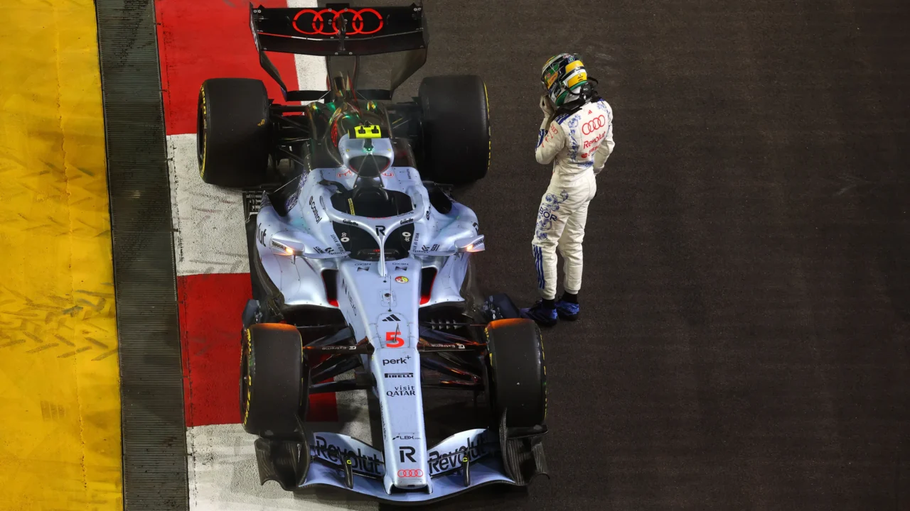Piloto de macacão branco com detalhes azuis ao lado de um carro de Fórmula 1 prateado parado na beira da pista, com a tampa do cockpit aberta