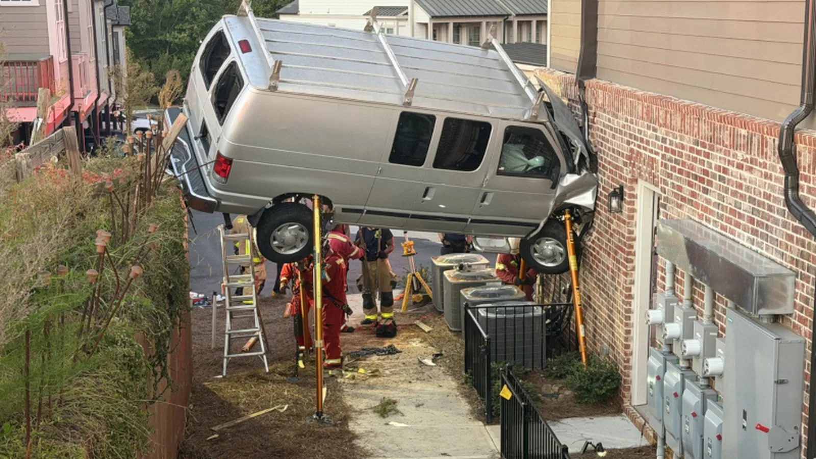 A van com a frente amassada contra o sobrado e as rodas no ar, apoiada em escoras, com bombeiros e uma escada embaixo, ainda com luz do dia